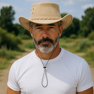May include: A tan-colored, wide-brimmed hat with a braided band and air vents. The hat has a curved brim and a chin strap. The person in the image is wearing a white t-shirt.