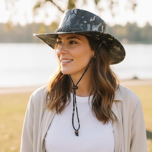 May include: A wide-brimmed hat with a camouflage pattern in shades of gray and black. The hat features a braided band around the crown and a chin strap. The hat is designed for outdoor activities and sun protection.