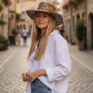 May include: A wide-brimmed hat with a snakeskin pattern in shades of brown and tan. The hat has a structured crown and a chin strap. The person is wearing a white button-down shirt and blue jeans.