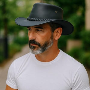 May include: A matte black fedora-style hat featuring a wide brim and a braided leather band around the crown. The hat includes a small metal ventilation eyelet on the side, providing a structured look with a smooth, solid finish throughout.