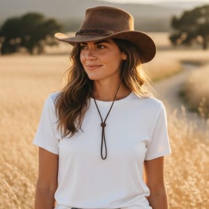 Op de afbeelding: Een bruine vilten fedora-hoed met een donkerbruine gevlochten leren band met metalen accenten en een bijpassend bruin kinbandje met een verstelbare sluiting, gecombineerd met een effen wit T-shirt met korte mouwen van glad katoen.