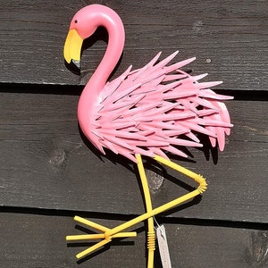 May include: Pink metal flamingo wall decoration with yellow legs and a yellow beak. The flamingo is standing on one leg with its wings spread out.