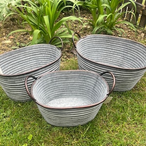 May include: Three galvanised metal oval planters with dark brown handles and rims. The planters are of varying sizes and sit on green grass. The ribbed texture of the metal is visible.