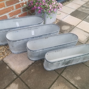 May include: Four galvanised metal oval planters in different sizes. The planters have a ribbed texture and a silver finish. They are arranged on a paved patio, with a brick wall and potted flowers in the background.