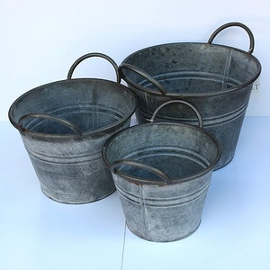 May include: Three galvanised metal buckets with black handles. The buckets are nested inside each other, with the largest bucket at the bottom and the smallest bucket at the top.