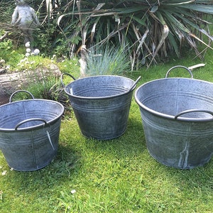 May include: Three galvanized metal buckets of different sizes with black handles are on a green lawn. The buckets have a classic, rustic design, suitable for gardening or decoration. The largest bucket is approximately 46 cm in diameter.