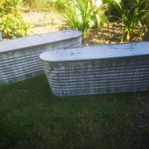 May include: Two oval-shaped, galvanised metal planters with a distressed finish. The tubs have a ribbed design and white accents. They are placed on grass, surrounded by greenery, creating a rustic garden aesthetic.