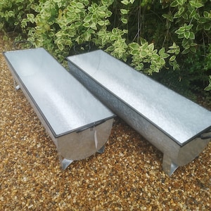 May include: Two galvanised metal troughs with black rims and handles. The troughs are sitting on a gravel surface.