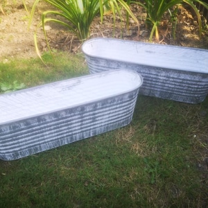 May include: Two oval-shaped metal planters with a distressed, weathered finish. The galvanized containers are painted white inside and sit on green grass. Ideal for outdoor gardening, these planters are a rustic addition.