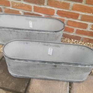 May include: Two oval galvanised metal planters with a ribbed design. The planters are grey and have a dark grey trim around the top edge. They are placed on a stone surface in front of a brick wall. Each planter has a small tag.