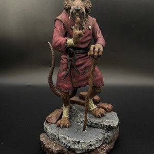 May include: A detailed, hand-painted figurine of Splinter, the rat sensei from the Teenage Mutant Ninja Turtles, in a red robe, holding a staff and standing on a rocky base.