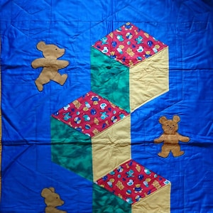 Könnte beinhalten: Eine blaue gesteppte Babydecke mit gelbem Rand. Die Decke zeigt drei gelbe und grüne Würfel mit einem rot-weiß-blauen Muster. Auf der Decke befinden sich drei braune Teddybären.