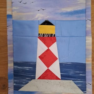 Peut inclure: Chemin de table en tissu matelassé avec un motif de phare. Le phare est blanc avec des losanges rouges et un sommet jaune. Le fond représente un ciel bleu, l'océan et des oiseaux en vol. Les bords du chemin de table ont un motif de vagues bleues et blanches.