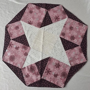 Op de afbeelding: Een achtkantige tafelrunner in roze en wit met een sneeuwvlokkenpatroon. De runner is gemaakt van stof en heeft een stervormig ontwerp in het midden.