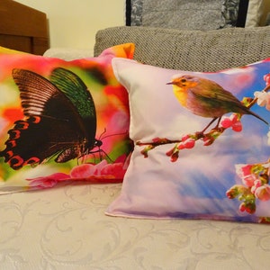 Può includere: Due cuscini decorativi con motivi floreali e animali colorati. Un cuscino presenta una farfalla nera e verde su uno sfondo rosa e giallo. L'altro cuscino presenta un piccolo uccello giallo appollaiato su un ramo con fiori rosa su uno sfondo di cielo blu.