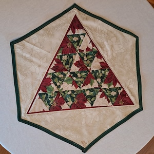 Pode incluir: Um caminho de mesa de Natal acolchoado com uma forma hexagonal única. O centro apresenta um triângulo de tecido de poinsétia, rodeado por um padrão floral creme e uma borda verde escura. O tecido de poinsétia tem flores vermelhas e folhas verdes.