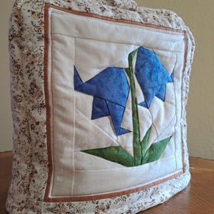 Puede incluir: Una funda de té acolchada con un diseño floral con flores azules y verdes. La funda tiene un borde de estampado floral marrón y blanco.