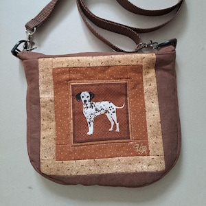 Puede incluir: Un bolso bandolera de tela marrón con un panel central cuadrado que muestra un perro dálmata impreso. El bolso tiene un borde acolchado en tonos beige y marrón con un patrón de puntos, una correa ajustable marrón y clips de metal negro.