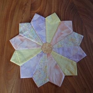 Könnte beinhalten: Ein handgefertigter Quiltblock mit einem Sternmuster in Rosa-, Gelb- und Lilatönen. Die Mitte des Blocks ist ein kleiner, runder, brauner Stoffkreis.