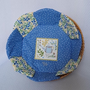 Könnte beinhalten: Ein runder Weidenkorb mit einem blau-weiß gepunkteten Stofffutter. Das Futter hat ein zentrales Quadrat mit einer Gießkannen-Illustration und floralen Akzenten. Stoffstücke mit floralem Muster in Gelb, Grün und Blau sind an den Rändern angenäht.