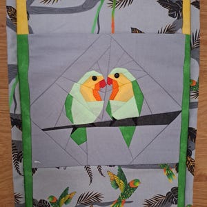 Puede incluir: Un artículo de tela gris con un diseño geométrico que presenta dos pájaros posados en una rama. Los pájaros son verdes, amarillos y naranjas. El fondo tiene un patrón de árboles y loros. El artículo está bordeado con ribetes verdes y amarillos.