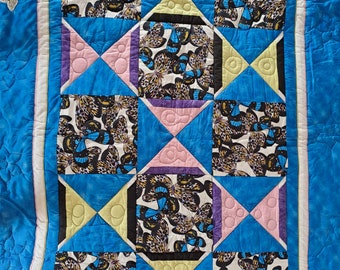 May include: A quilt featuring a repeating pattern of butterfly-print fabric squares alternating with blue, yellow, and pink triangular geometric shapes. The quilt has a bright blue border with small butterfly appliques and white stitching.