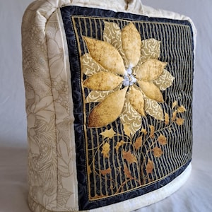 Könnte beinhalten: Ein gesteppter Stoffbezug für eine Kaffeemaschine oder ein anderes Küchengerät. Der Bezug ist cremefarben mit einem schwarzen und goldenen Blumendesign. Das Design zeigt eine große Weihnachtsstern mit einem Strassstein in der Mitte.