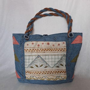Puede incluir: Un bolso tote de mezclilla azul con un asa de cuerda marrón y un diseño de patchwork con un patrón floral y formas geométricas. El bolso tiene dos cierres de botón.
