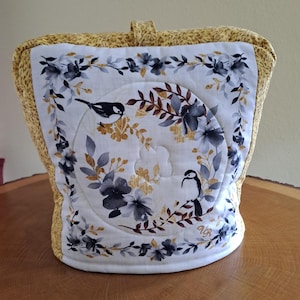 Op de afbeelding: Een gewatteerde theemuts met een wit middenpaneel met een bloemen- en vogelontwerp in grijs, goud en bruin. De randen zijn afgezet met een geel bloemmotief en hebben een bijpassende handgreep.