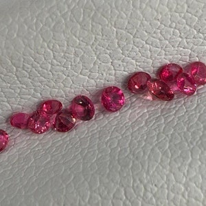 May include: A scattering of small, oval-shaped, bright pink gemstones on a white, textured surface. The polished gemstones reflect light, highlighting their vibrant colour and clarity. The arrangement suggests a display of loose gemstones.