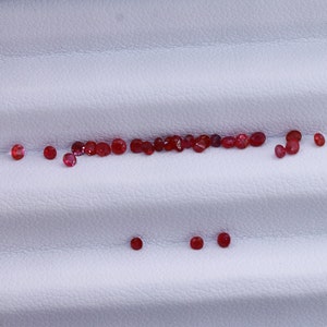 Può includere: Una collezione di piccole pietre preziose rosse disposte in linea su una superficie bianca testurizzata.