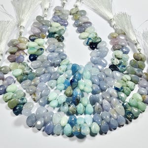 May include: Strands of gemstone beads in shades of blue, green, and purple, with white tassel accents. The beads are teardrop-shaped and arranged in a cascading pattern, creating a gradient effect. The gemstones are polished and have a natural, organic appearance.