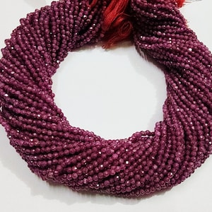 Op de afbeelding: Een streng gefacetteerde, diep bordeauxrode kralen. De kralen zijn strak aan elkaar geregen en vormen een gedraaide, touwachtige vorm. De kleur is een rijk, donkerrood met een lichte glans, wat wijst op een edelsteenmateriaal. Een klein bundeltje rode draden is bovenaan zichtbaar.
