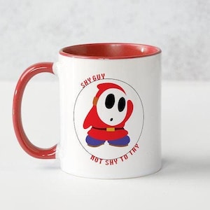 May include: A white ceramic mug with a red rim and handle. The mug features a cartoon image of a Shy Guy from the Super Mario Bros. video game series. The image is surrounded by a circle with the text "Shy Guy Not Shy To Try".