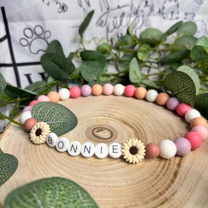 Peut inclure: Un collier pour chien avec le nom "Bonnie" épelé avec des perles blanches. Le collier présente un dégradé de perles roses, pêche et blanches, avec deux petits accents de tournesol. Le collier est présenté sur une surface en bois.