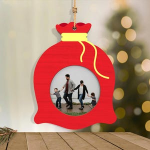 May include: A red wooden Christmas ornament shaped like a gift bag, with a yellow drawstring and a circular photo frame. The frame contains a family photo. The ornament hangs from a natural fiber cord.