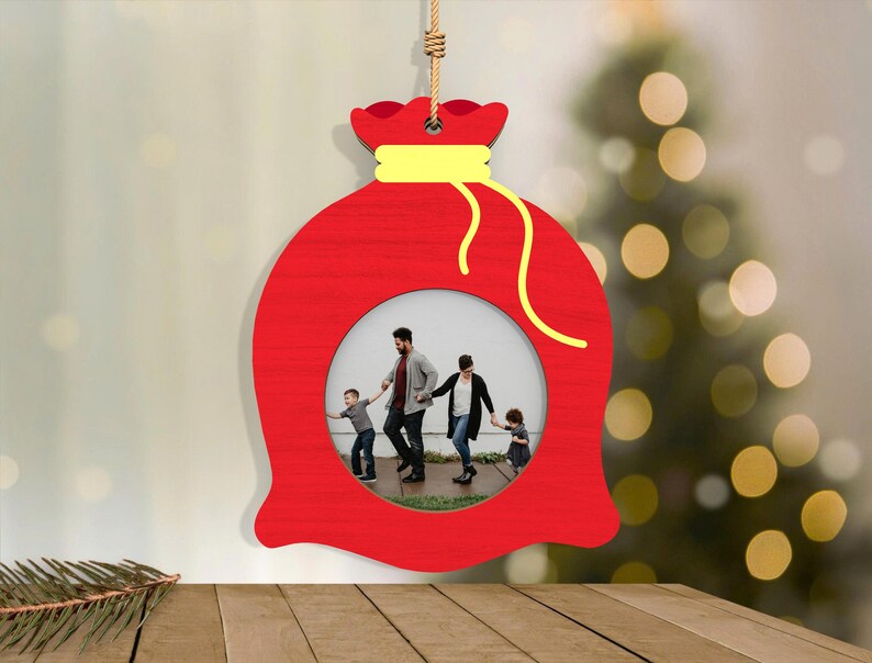 May include: A red wooden Christmas ornament shaped like a gift bag, with a yellow drawstring and a circular photo frame. The frame contains a family photo. The ornament hangs from a natural fiber cord.
