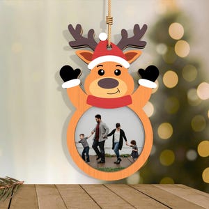 May include: A wooden Christmas ornament shaped like a reindeer with a photo frame. The reindeer has a red and white Santa hat, black mittens, and a red scarf. The frame holds a family photo. The ornament hangs from a natural twine.