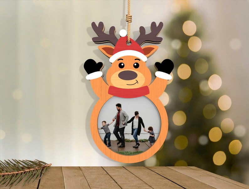 May include: A wooden Christmas ornament shaped like a reindeer with a photo frame. The reindeer has a red and white Santa hat, black mittens, and a red scarf. The frame holds a family photo. The ornament hangs from a natural twine.