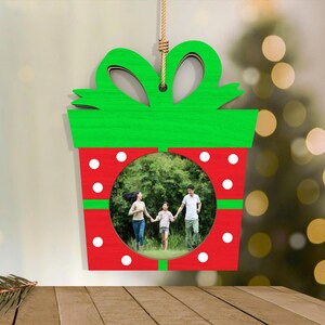 May include: A Christmas ornament shaped like a gift box, with a green bow and red body. The ornament has a circular opening displaying a family photo. White polka dots decorate the red section, and it hangs from a natural rope.
