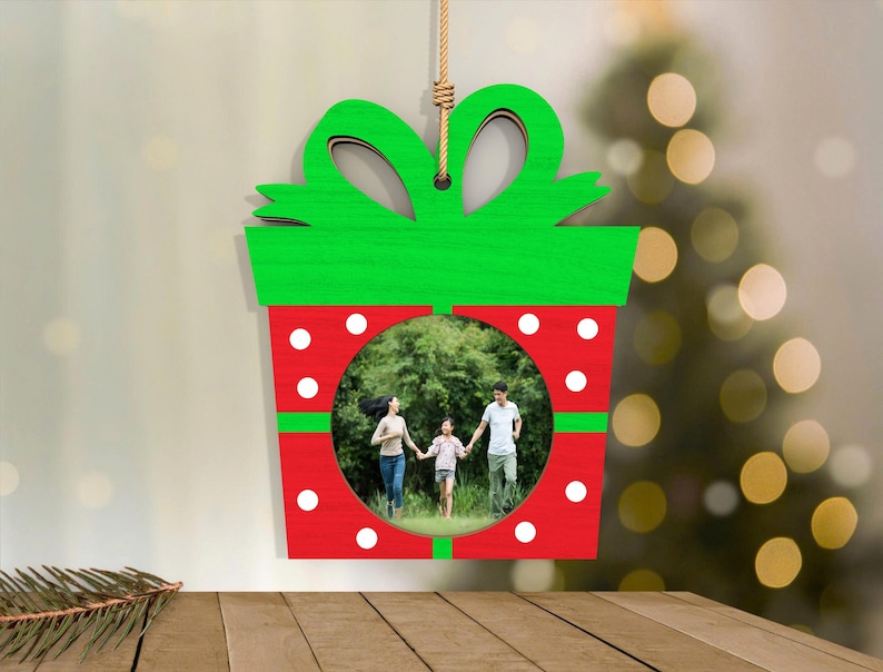 May include: A Christmas ornament shaped like a gift box, with a green bow and red body. The ornament has a circular opening displaying a family photo. White polka dots decorate the red section, and it hangs from a natural rope.