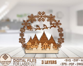 May include: A wooden Christmas-themed decoration featuring a winter village scene with houses, trees, and snowmen. A snowflake-shaped wreath surrounds the scene, with Santa's sleigh and reindeer. The base is light-colored wood, and the design is laser cut. The text "DIGITAL FILES FOR LASER CUT" is visible.
