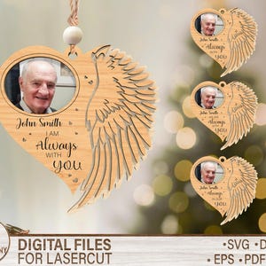 May include: Wooden heart-shaped memorial ornament with a photo of a man, angel wing design, and the text "John Smith I am always with you." Includes a string for hanging. Additional ornaments are visible in the background.