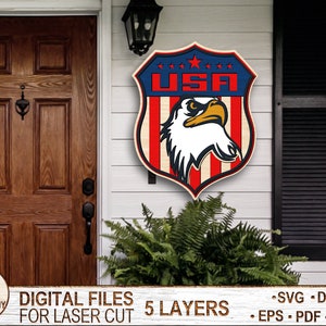 May include: A wooden cutout of a shield with the word "USA" and an eagle inside. The shield is red, white, and blue. The eagle is white with a black beak and eyes. The cutout is on a white wooden door with a brown wooden door to the left. The cutout is for laser cutting and comes in 5 layers.