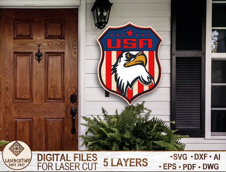 May include: A wooden cutout of a shield with the word "USA" and an eagle inside. The shield is red, white, and blue. The eagle is white with a black beak and eyes. The cutout is on a white wooden door with a brown wooden door to the left. The cutout is for laser cutting and comes in 5 layers.