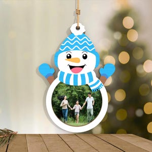 May include: A snowman-shaped photo frame with a blue hat, mittens, and scarf. The frame holds a family photo. The snowman is hanging by a string, and the background is blurred with a Christmas tree.