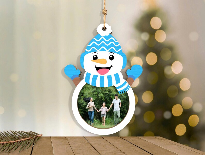 May include: A snowman-shaped photo frame with a blue hat, mittens, and scarf. The frame holds a family photo. The snowman is hanging by a string, and the background is blurred with a Christmas tree.