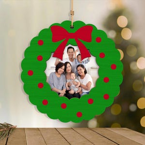 May include: A green wooden Christmas wreath ornament with a red bow and red dot accents. The center displays a family photo. The ornament hangs from a natural twine string, perfect for holiday decor.