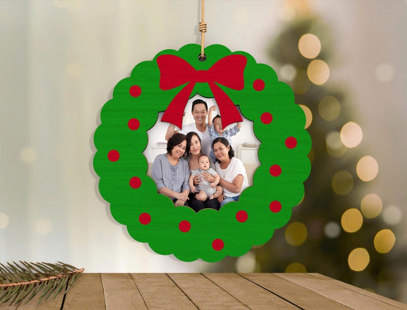 May include: A green wooden Christmas wreath ornament with a red bow and red dot accents. The center displays a family photo. The ornament hangs from a natural twine string, perfect for holiday decor.
