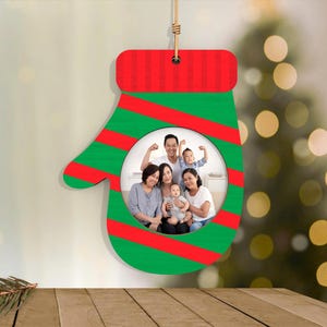 May include: A Christmas ornament shaped like a mitten, with a family photo in the center. The mitten is green with red stripes and a red top. A string is attached for hanging. The background is blurred with a Christmas tree.